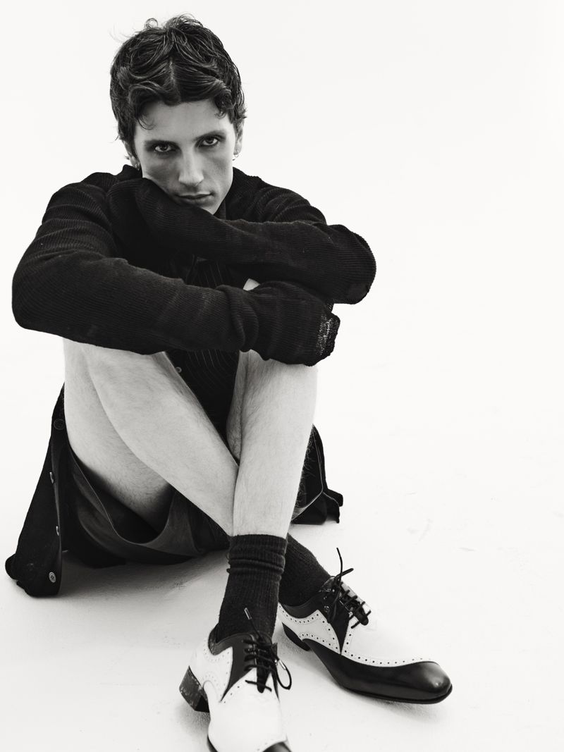 Young man in dark jacket and socks sits on the floor with legs crossed and black and white dress shoes on a bright white background.