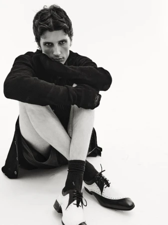 Young man in dark jacket and socks sits on the floor with legs crossed and black and white dress shoes on a bright white background.