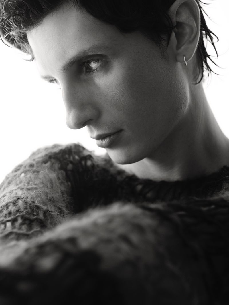 Striking black and white portrait of a person with short, dark hair and an earring, wearing a textured sweater, against a bright backdrop.