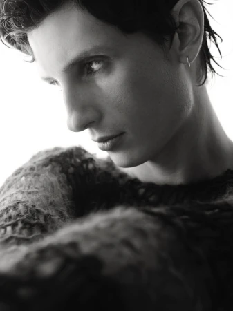 Striking black and white portrait of a person with short, dark hair and an earring, wearing a textured sweater, against a bright backdrop.