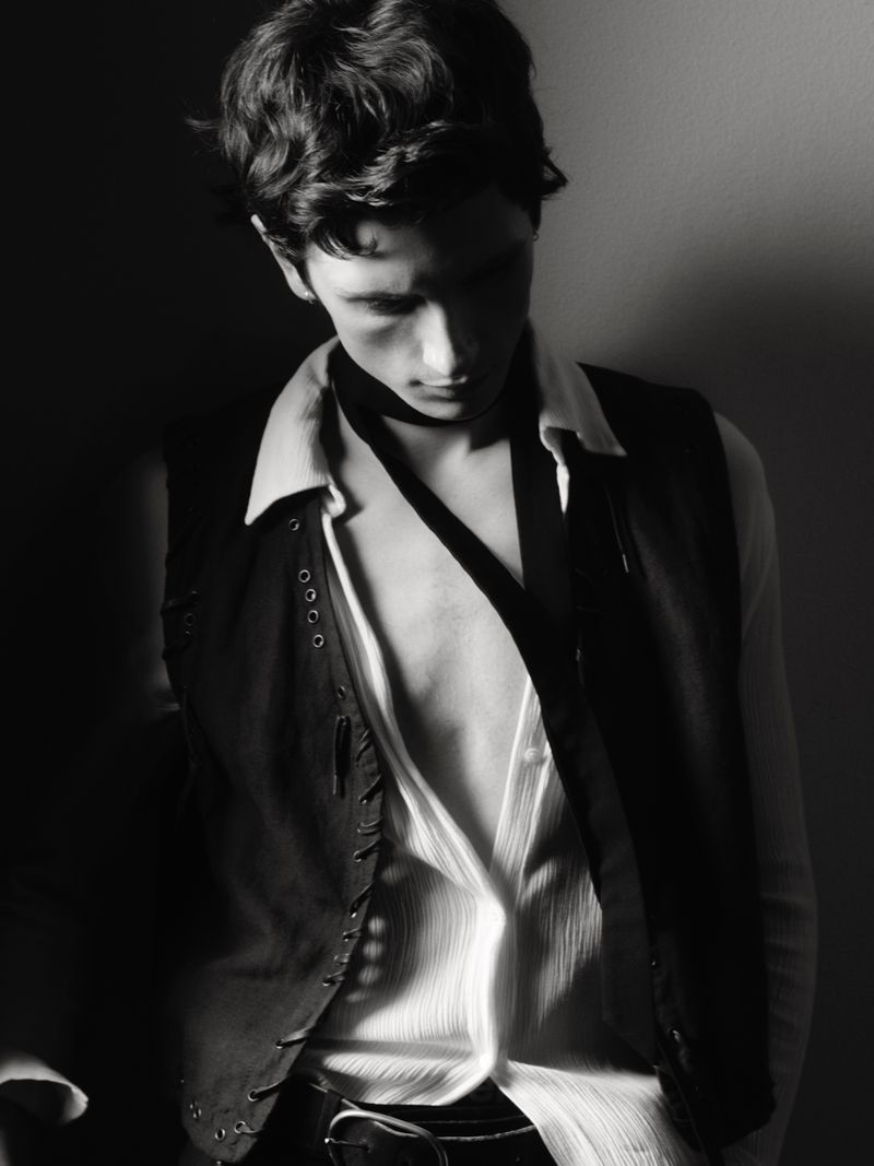 Monochrome portrait of a young man in a vest and open white shirt, looking down with a shadow across his face in a dim setting.