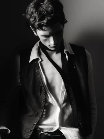 Monochrome portrait of a young man in a vest and open white shirt, looking down with a shadow across his face in a dim setting.