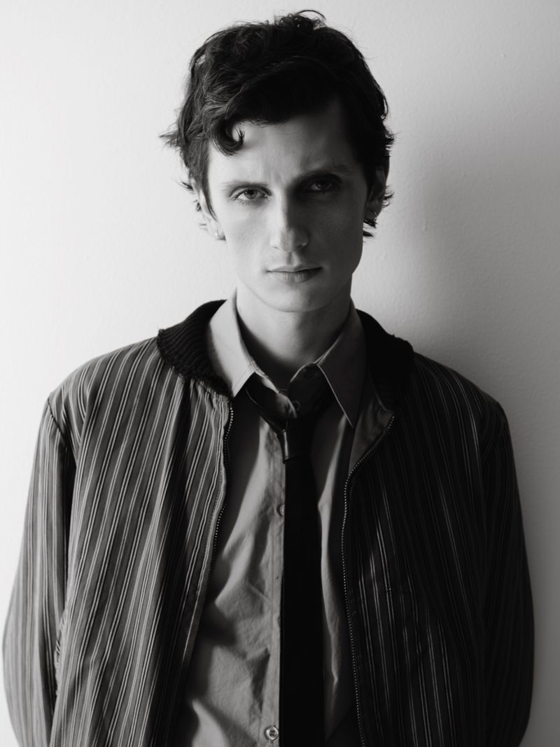 Black and white portrait of a young adult man in a striped jacket and open button-down shirt with a tie, against a plain background.