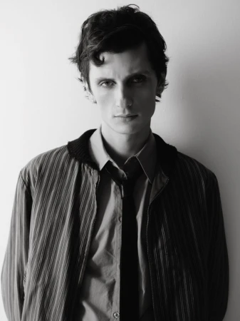 Black and white portrait of a young adult man in a striped jacket and open button-down shirt with a tie, against a plain background.