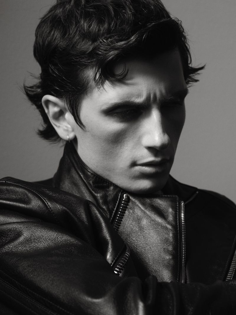 Monochrome portrait of a dark-haired man in a leather jacket against a plain background, captured in a close-up shot with a serious expression.