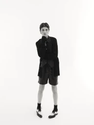 Model Jack Stewart standing in a layered black cardigan, shorts, and two-tone dress shoes against a white seamless background.