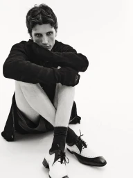 Model Jack Stewart seated cross-legged on a white floor wearing a black long-sleeve top and two-tone spectator shoes.