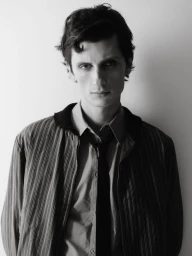 Model Jack Stewart wearing a striped zip-up jacket, collared shirt, and slim tie against a plain studio background.
