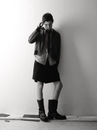 Model Jack Stewart posing against a white wall wearing a jacket, shirt, tie, tailored shorts, and black leather harness boots.