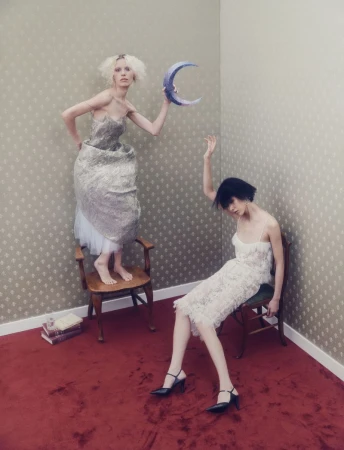 Two women in dresses pose in a room with patterned wallpaper and a red carpet. One is standing on a chair, holding a crescent moon.