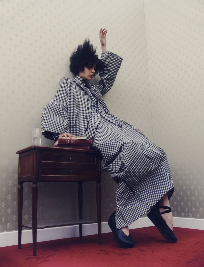Young adult poses in patterned jacket and dress leaning on a wooden side table with books, glass of water indoors, red carpet.