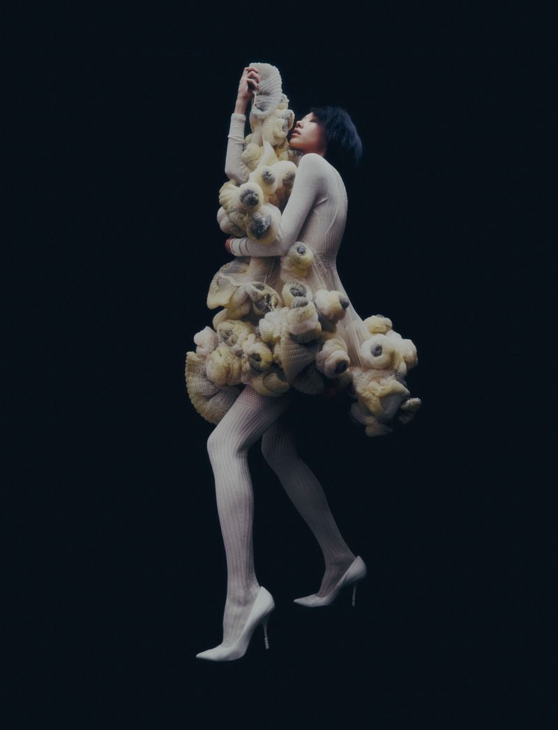 A woman in a sculptural cream-colored dress stands against a dark background in this striking fashion photograph with full-body view.