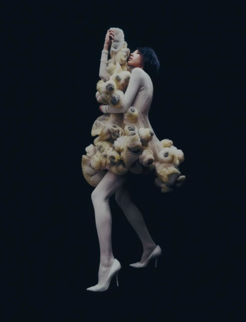 A woman in a sculptural cream-colored dress stands against a dark background in this striking fashion photograph with full-body view.
