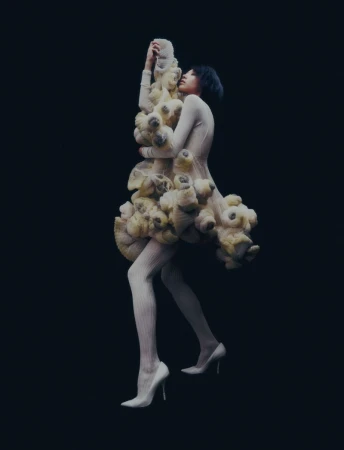 A woman in a sculptural cream-colored dress stands against a dark background in this striking fashion photograph with full-body view.
