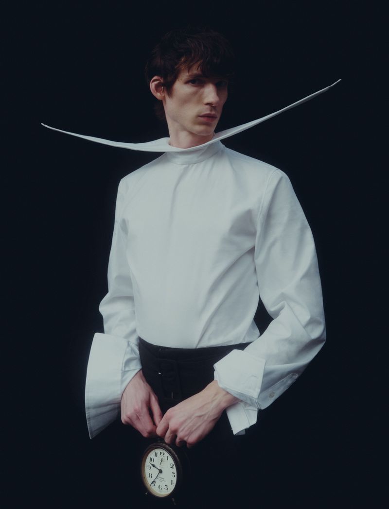 Fashionable adult male in a stark white shirt with a wide collar, posing against a black backdrop while holding an antique clock.