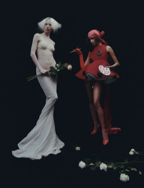Two fashion models pose in a studio setting; one is wearing a white gown and holding roses, and the other wears a red artist-themed costume.
