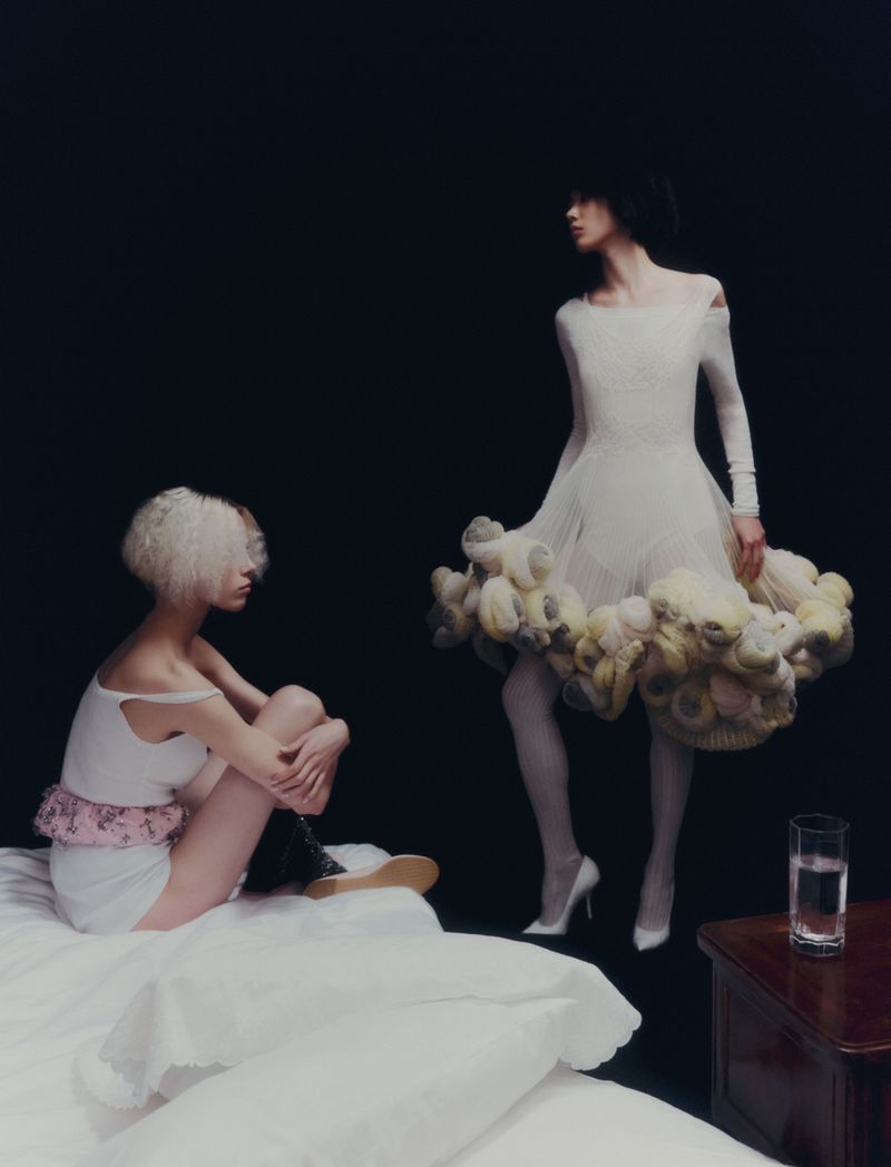 Two women in light-colored dresses pose in a studio. One is sitting on a bed. The other stands near a glass of water on a wood table.