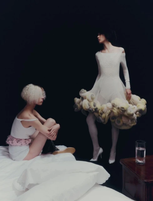 Two women in light-colored dresses pose in a studio. One is sitting on a bed. The other stands near a glass of water on a wood table.