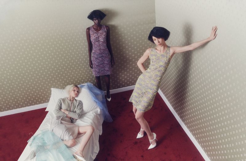 Three women in fashionable dresses pose in an indoor setting with patterned walls and red carpet; one woman lies on a bed.
