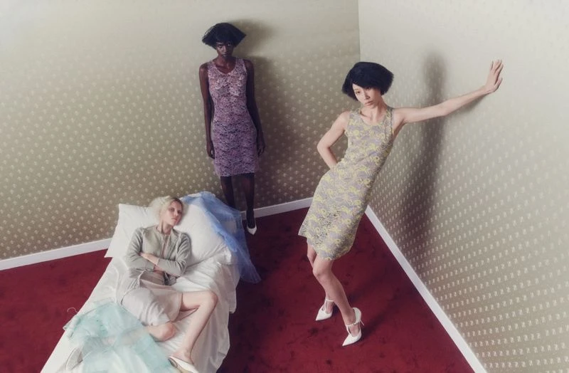 Three women in fashionable dresses pose in an indoor setting with patterned walls and red carpet; one woman lies on a bed.