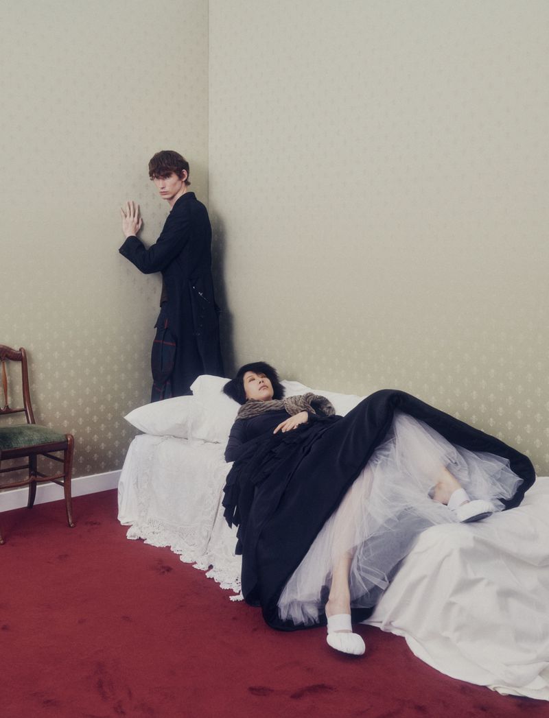Indoor fashion scene with two people: a man leans against the wall, and a woman lies on a bed with a long black dress and white shoes.