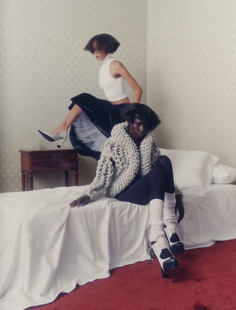 Two Black female fashion models pose indoors, one seated on a bed in a beige sweater and the other on a table, wearing a white top and skirt.