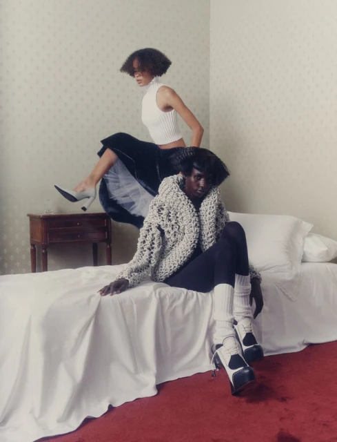 Two Black female fashion models pose indoors, one seated on a bed in a beige sweater and the other on a table, wearing a white top and skirt.