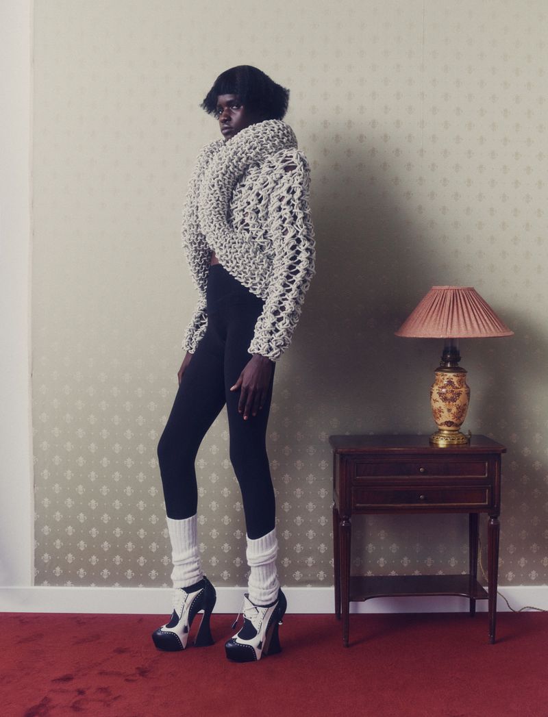 Fashionable woman posing in a patterned sweater, black leggings, leg warmers, and platform heels near a table lamp indoors.