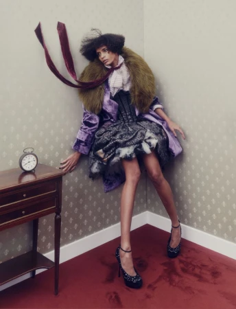 Editorial fashion shot: a slender woman in haute couture posing in the corner of a room next to a wooden table with an alarm clock.