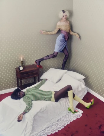 Fashionable women in an indoor setting. One is lying on a bed with green knitwear and tall socks, while the other steps down.