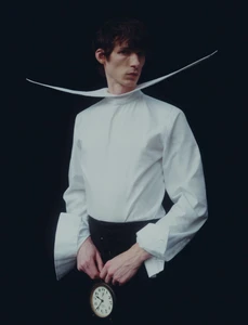 A man in an avant-garde white shirt with an exaggerated curved collar holding an analog clock against a black backdrop.