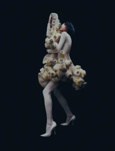 Model posing in an avant-garde cream textured sculptural dress, ribbed tights, and white pointed high heels against a black studio backdrop.