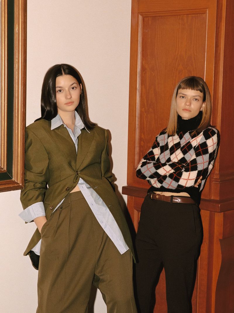 Two young women pose indoors in fashion-forward outfits. One wears an oversized blazer and the other an argyle sweater.