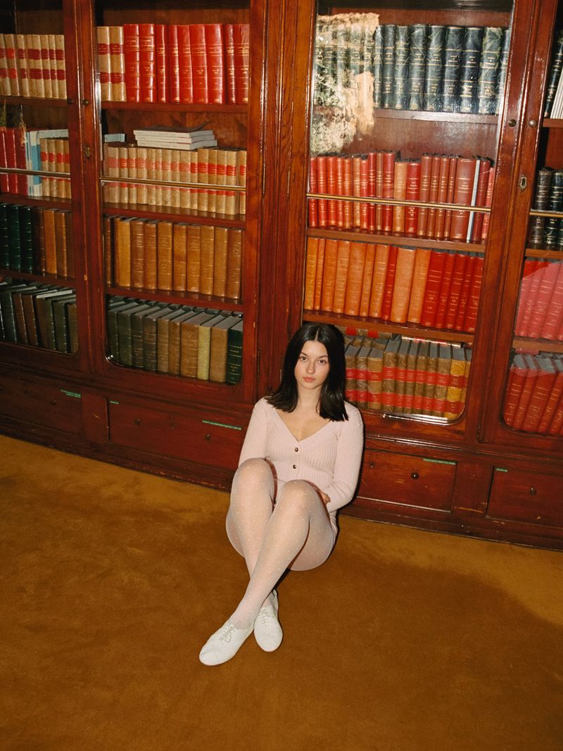 Brunette woman in pink outfit sits on brown carpet near a large wooden bookshelf filled with many books in an indoor setting.