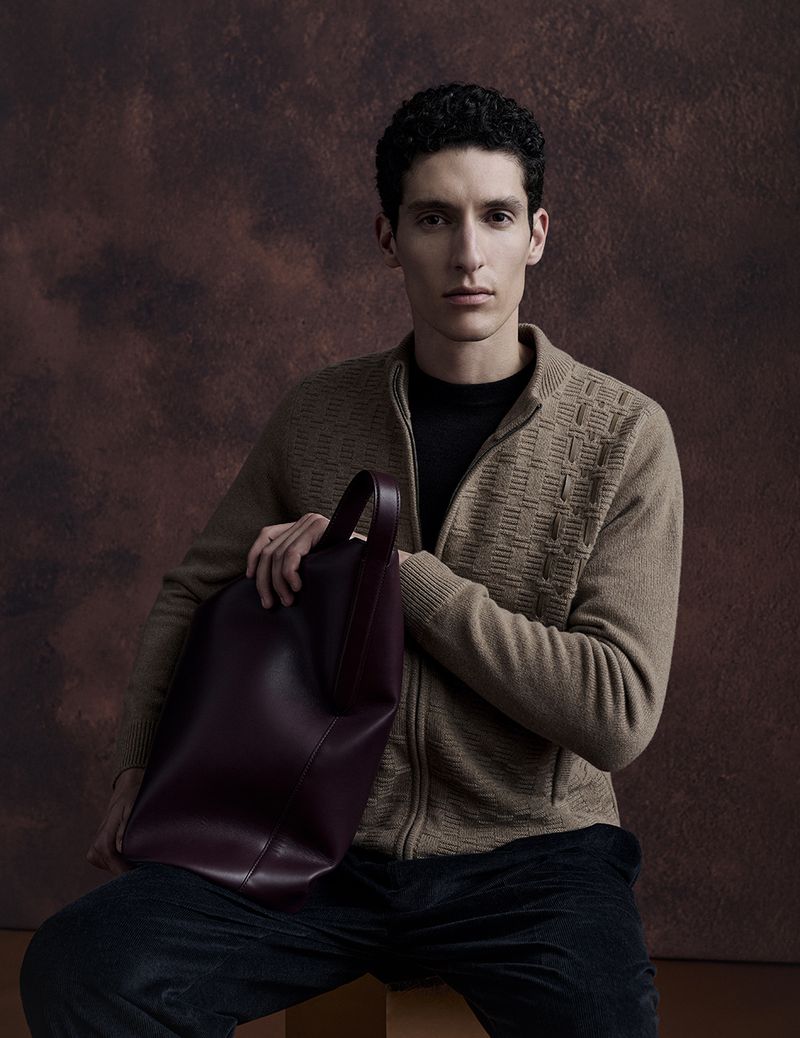 An adult male with curly hair poses in a brown sweater and black pants, holding a large burgundy leather tote bag against a brown background.