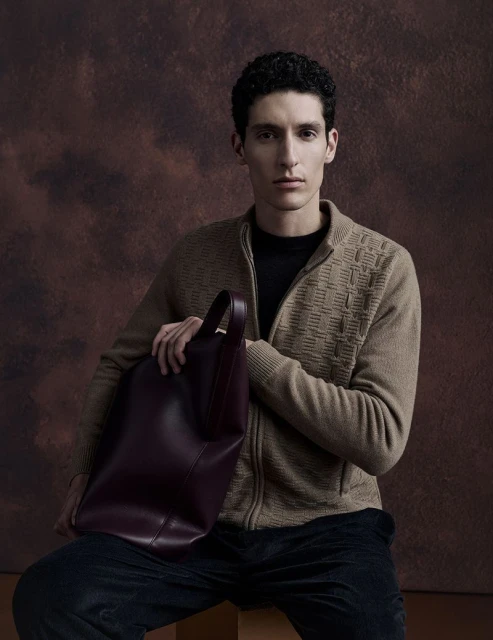 An adult male with curly hair poses in a brown sweater and black pants, holding a large burgundy leather tote bag against a brown background.