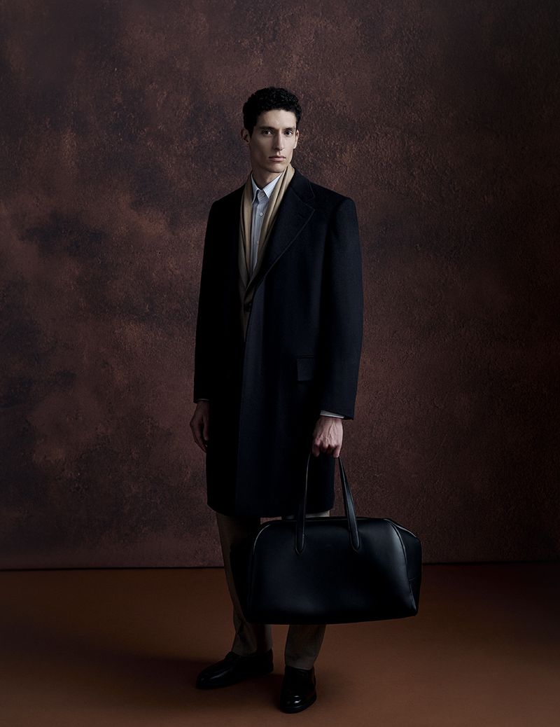 Fashionable adult male model strikes a pose in a studio, wearing a black overcoat, holding a dark leather bag, against a brown mottled backdrop.