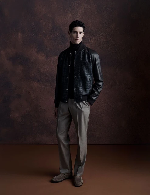 A full shot of a man in a black jacket and light-colored pants posing against a textured brown background in a studio setting.