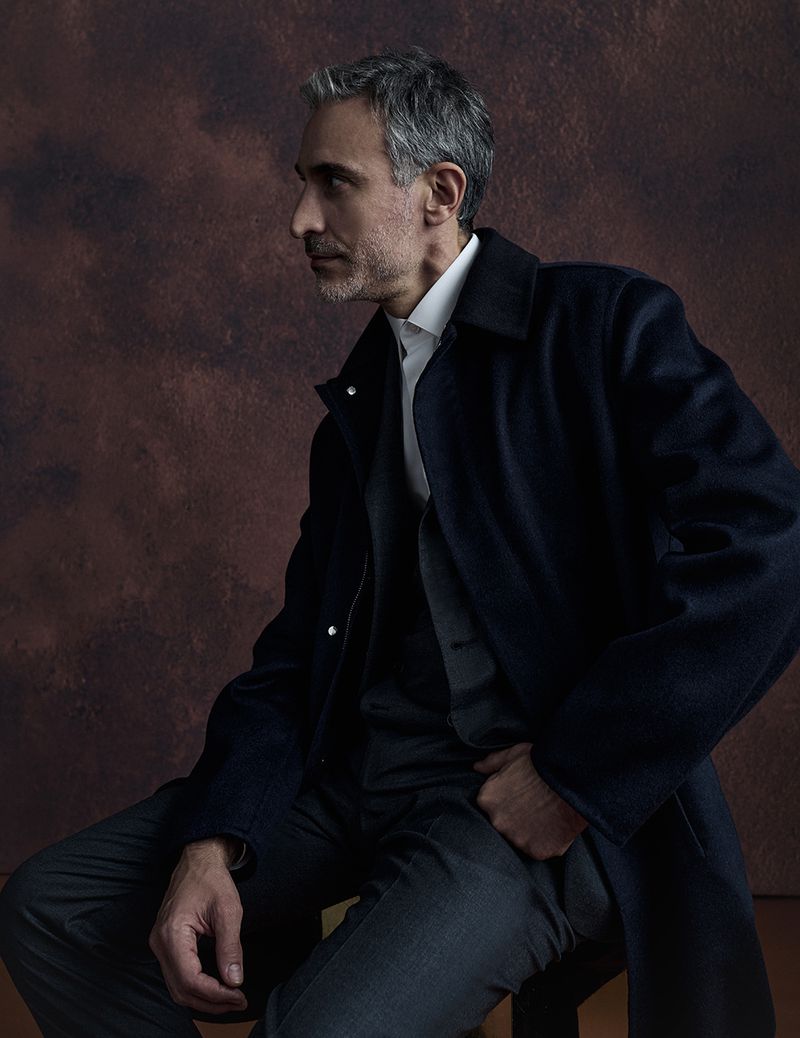 Portrait of an adult male with gray hair wearing a dark overcoat, vest, collared shirt and trousers while sitting on a stool.