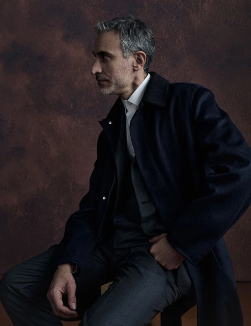 Portrait of an adult male with gray hair wearing a dark overcoat, vest, collared shirt and trousers while sitting on a stool.