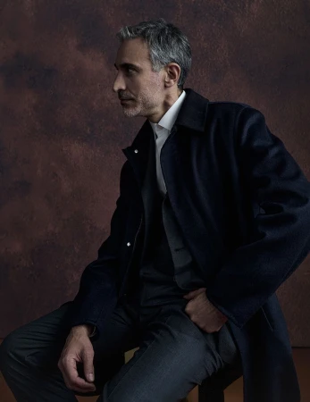 Portrait of an adult male with gray hair wearing a dark overcoat, vest, collared shirt and trousers while sitting on a stool.