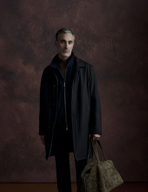 A man in a coat is holding a bag. He is standing indoors with a neutral expression and dark clothing in front of a brown background.