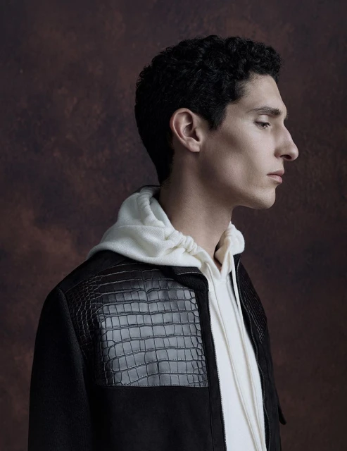 Profile portrait of a young adult man in a black jacket with reptile skin detail and white hoodie, against a mottled brown backdrop.