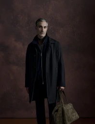Man in a dark overcoat holding an olive green suede duffle bag against a textured dark brown studio backdrop.
