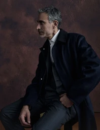 Middle-aged man with salt-and-pepper hair wearing a navy coat over a dark suit and white shirt against a textured brown backdrop.