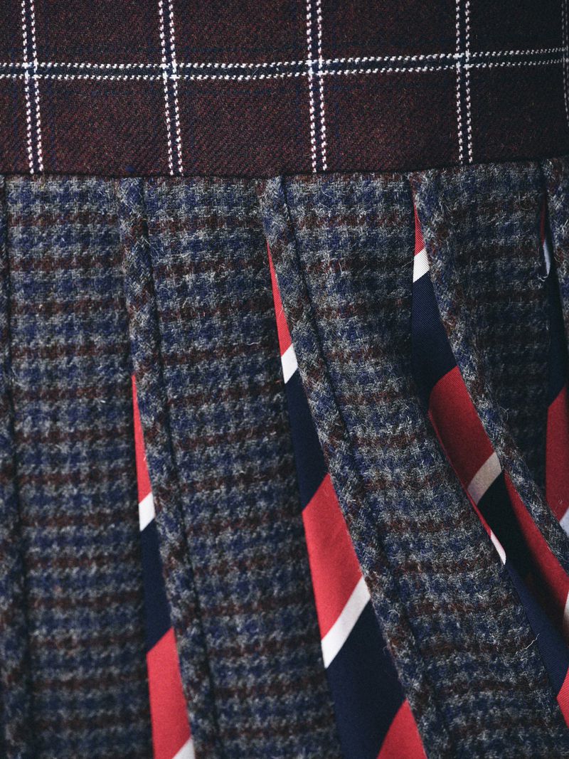 Close-up of a pleated skirt featuring a brown and white plaid waistband, a navy and brown houndstooth pattern, and striped red, white and blue panels.