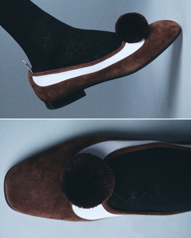 Elevated shoe-fashion image. Brown suede loafer with white trim and dark pom-pom is shown worn with black sock. Split view.