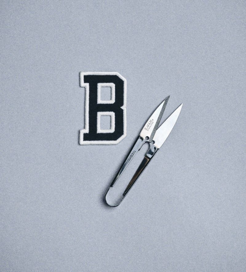 An embroidered varsity letter B patch with black trim is lying next to sewing scissors on a solid grey background in a studio setting.