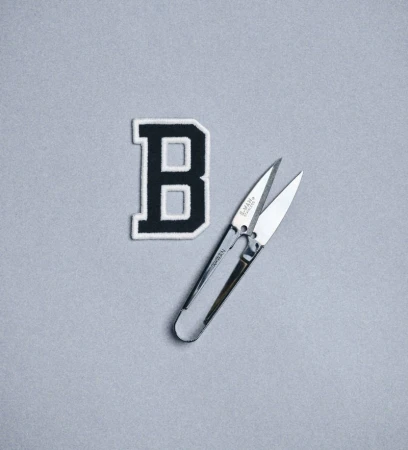 An embroidered varsity letter B patch with black trim is lying next to sewing scissors on a solid grey background in a studio setting.