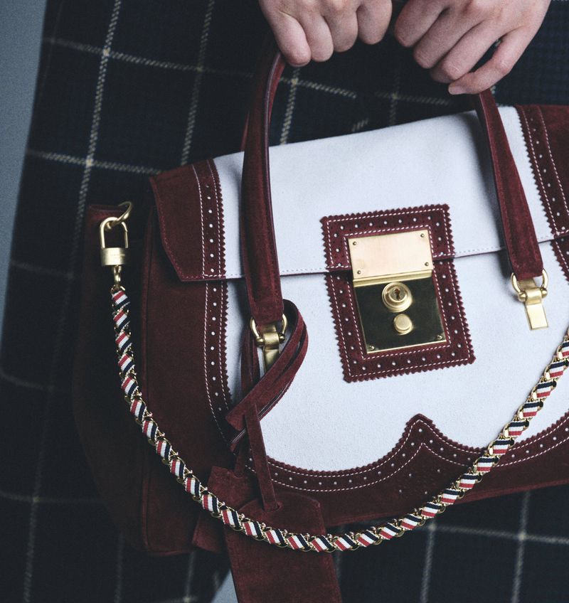 White and burgundy leather satchel handbag with gold hardware held by a person in a plaid skirt, studio shot.